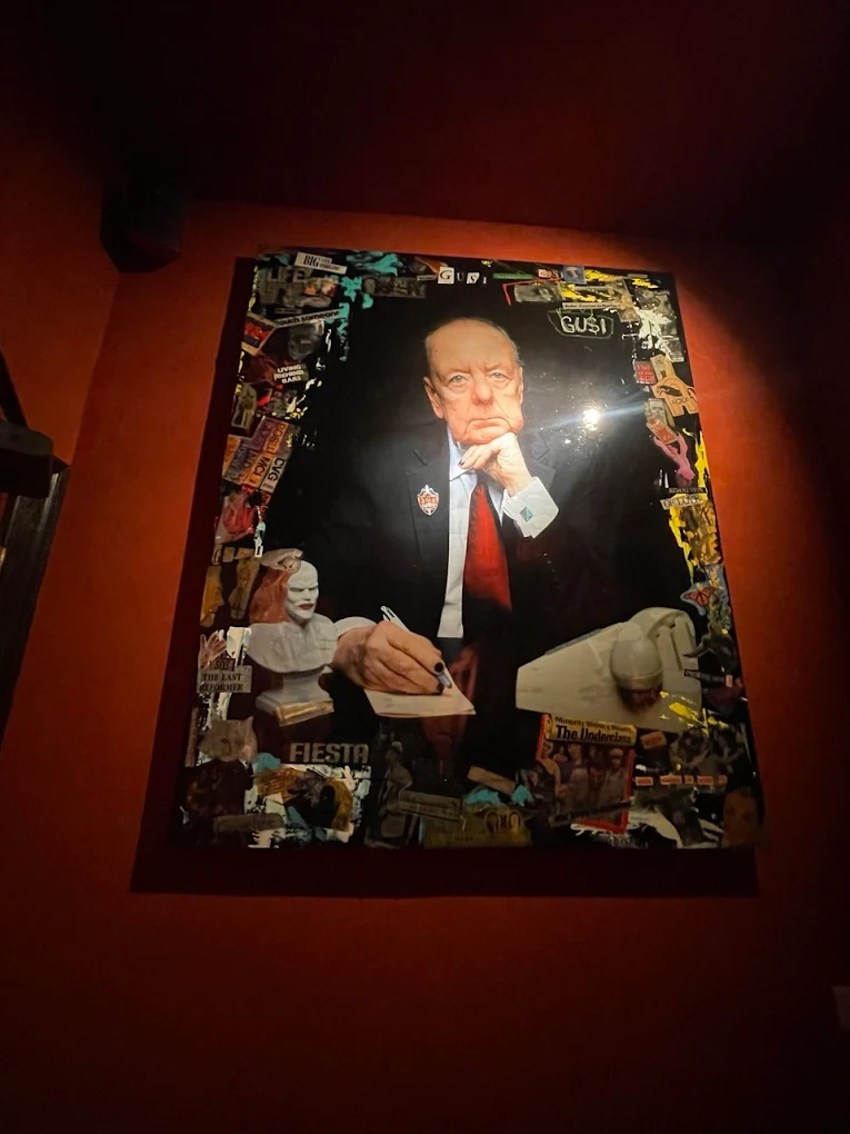 An original commissioned mixed-media portrait at GUSI Restaurant — an older gentleman seated and writing, set against a dense collage of magazine clippings, hung on a deep red wall