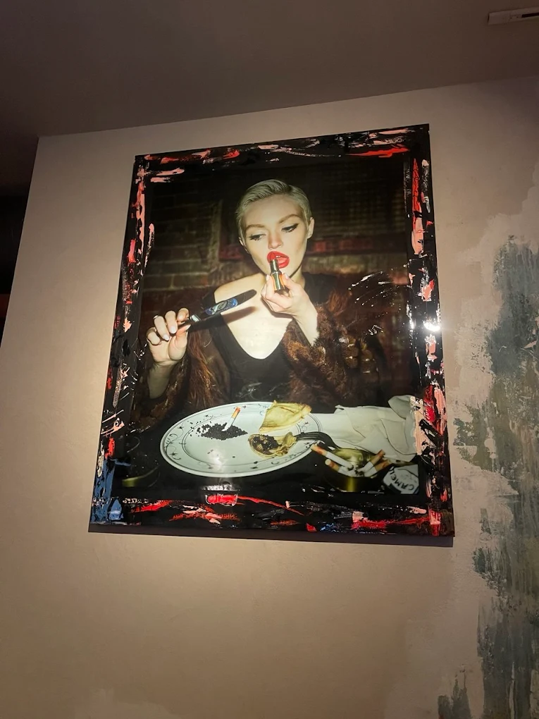 An original commissioned artwork at GUSI Restaurant — a large painterly portrait of a woman in fur applying red lipstick at a dinner table, framed by an expressionist red and black border, displayed on a textured plaster wall
